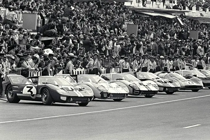 Row of Ford GT40 race cars lined up on the Le Mans starting grid in the 1960s with crowds watching from the stands.