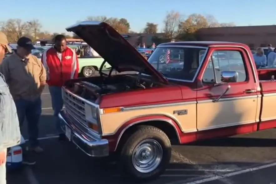 Two-tone red-and-tan 1980s Ford F-150 with hood open at a car show, surrounded by spectators.