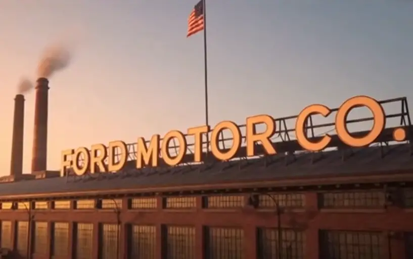 Ford Motor Company factory building at sunset with smokestacks and American flag flying above the roof.